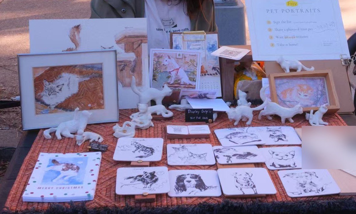 Market table with framed pet paintings, white clay cats and ink pet portraits on coasters, with a Free Pet Portraits sign