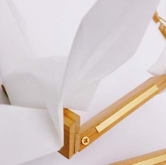 Close-up of folded white paper crane wings meeting a carved wood arm with a brass inlay and screw