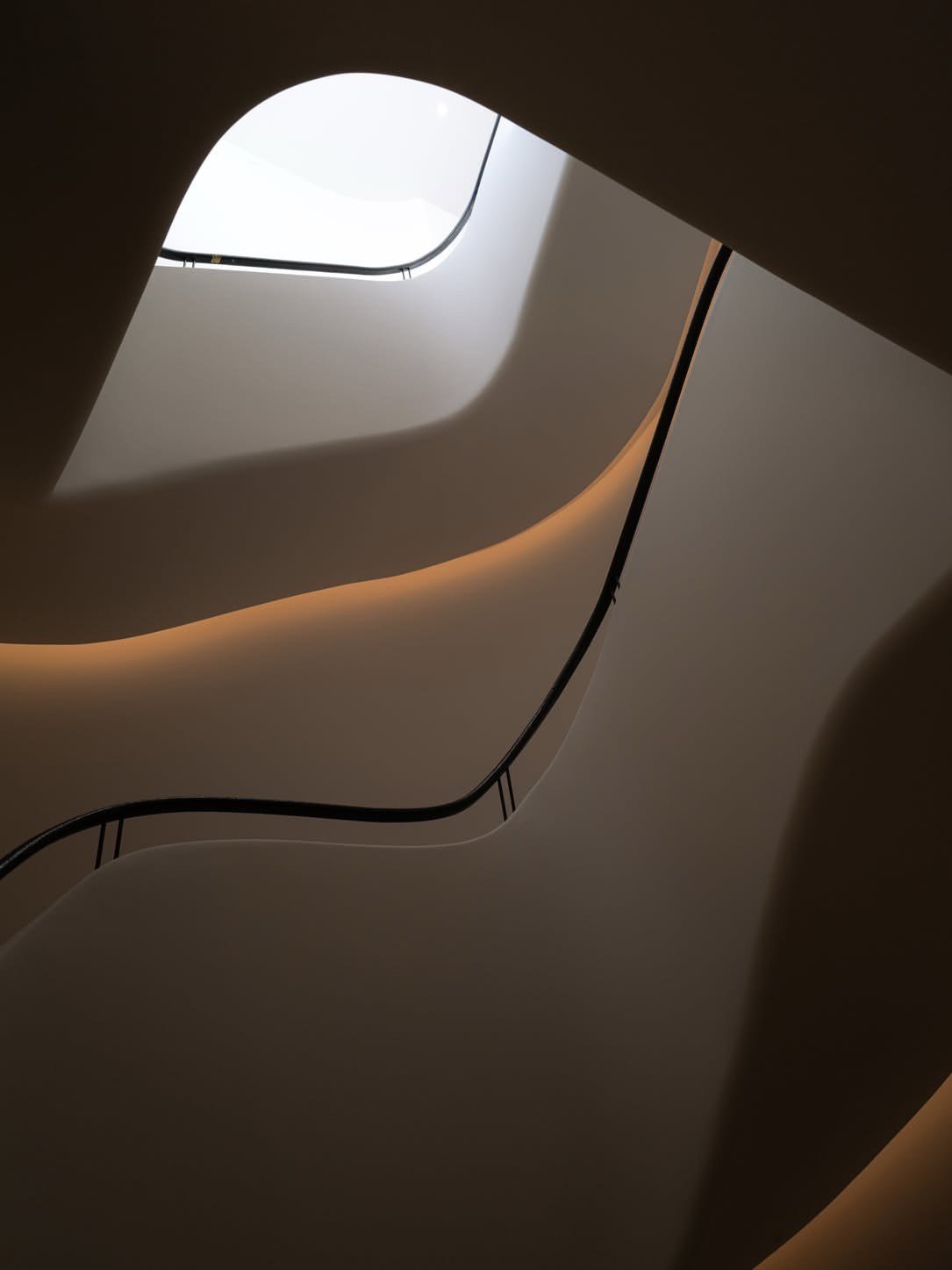 Looking up a sculptural white stairwell toward a skylight, a soft glow grazing the curved underside